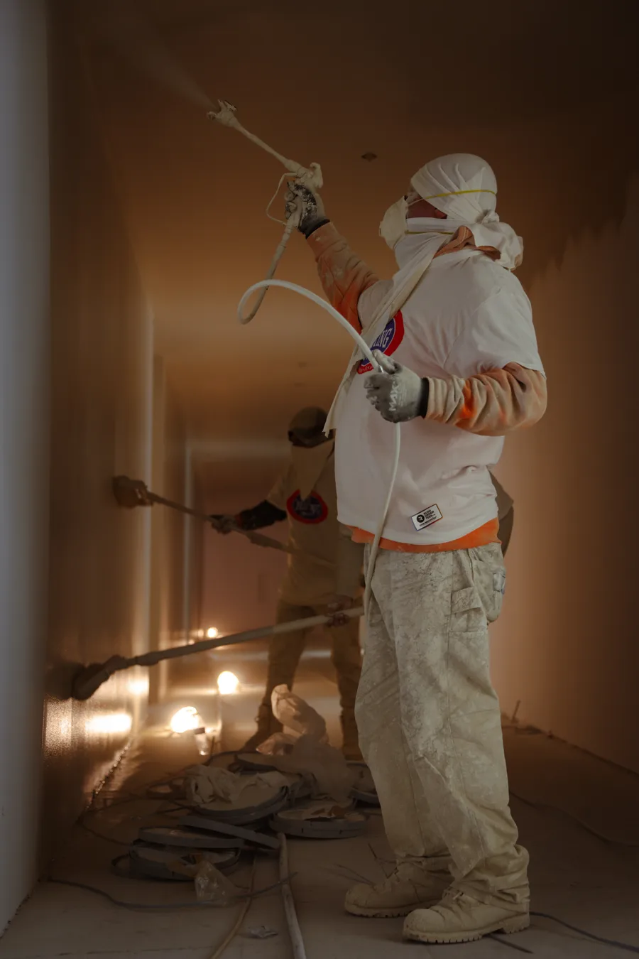 An RC Painting & Sons painter spraying a ceiling with an extension-pole gun, hooded and masked against the overspray in a white RC-logo shirt, with a second painter working further down the corridor behind.
