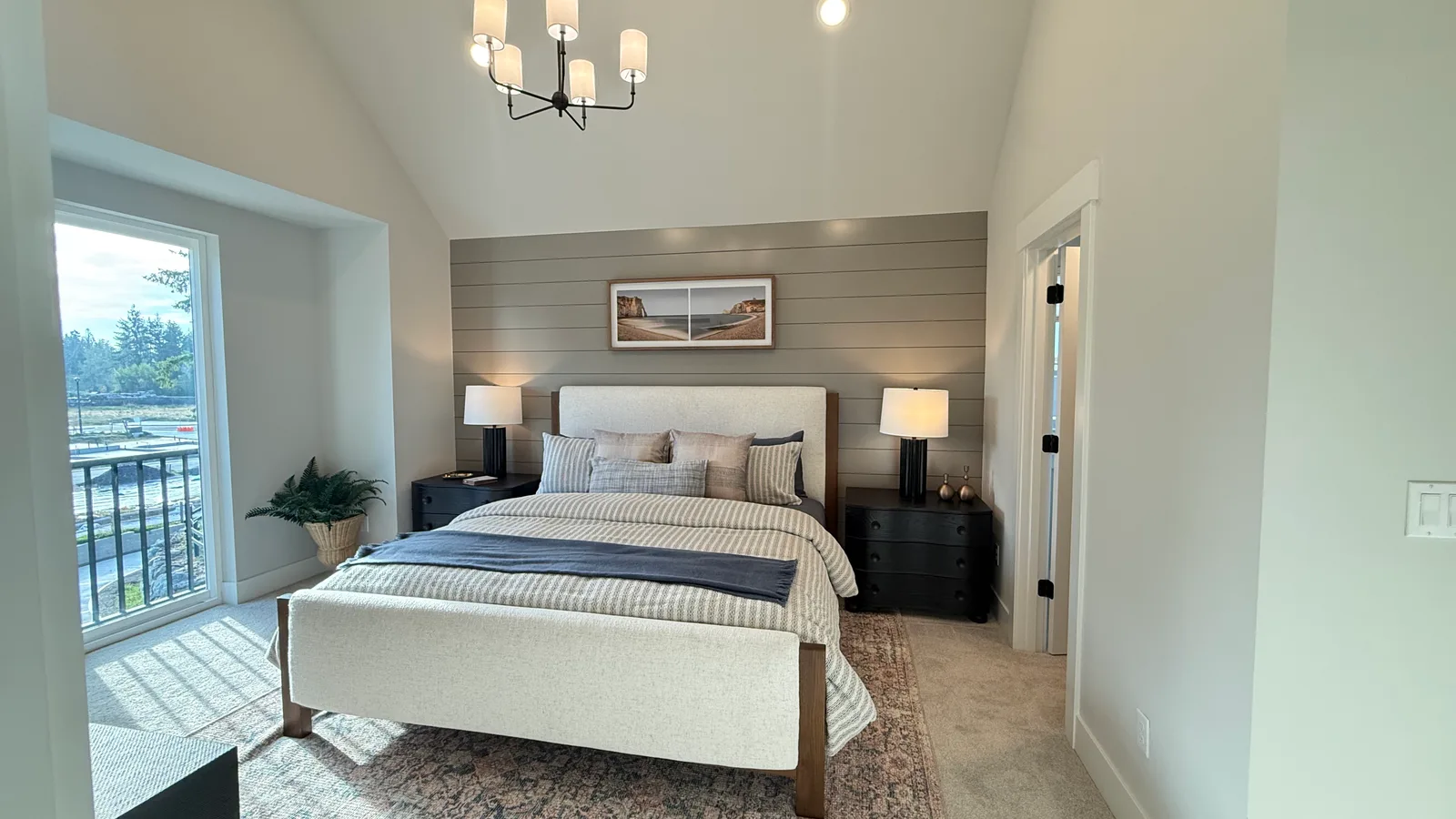 A primary bedroom at Brownstone in Sammamish: a vaulted white ceiling with a black-and-white chandelier, a warm grey shiplap accent wall behind an upholstered bed, and a glass door opening onto a black railed balcony.