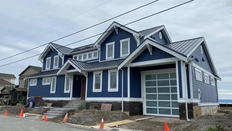 A two-storey custom home on Camano Island in deep navy shingle siding with crisp white trim, gables and a standing-seam metal roof, a frosted glass garage door at one end, and the water just visible past the yard.