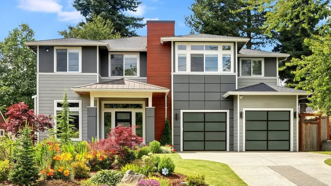 A two-storey modern custom home in grey lap siding and grey panels, a cedar-clad chimney rising through the centre, a standing-seam copper roof over the entry, two dark glass-panel garage doors, and a planted front garden of Japanese maples and daylilies.