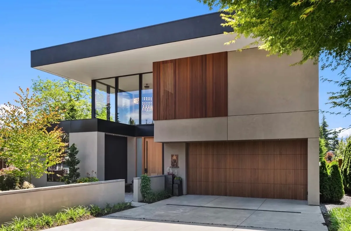 A modern two-storey custom home in Kirkland: smooth grey plaster walls under a deep flat roof overhang, a vertical cedar-slat panel above a matching cedar garage door, floor-to-ceiling glass at the stair, and a concrete drive between low planted walls.