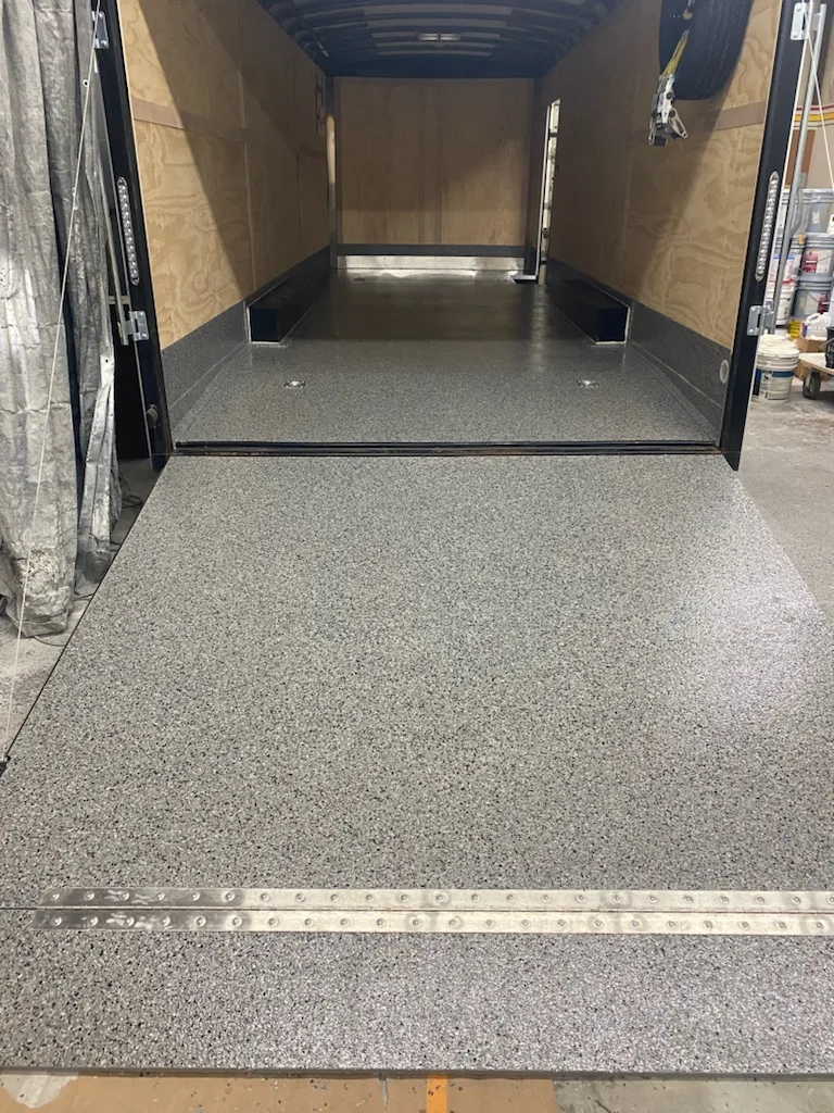 An enclosed trailer seen from the ramp: grey flake coating over the ramp and the floor running back between plywood walls, with a steel threshold strip across the ramp.