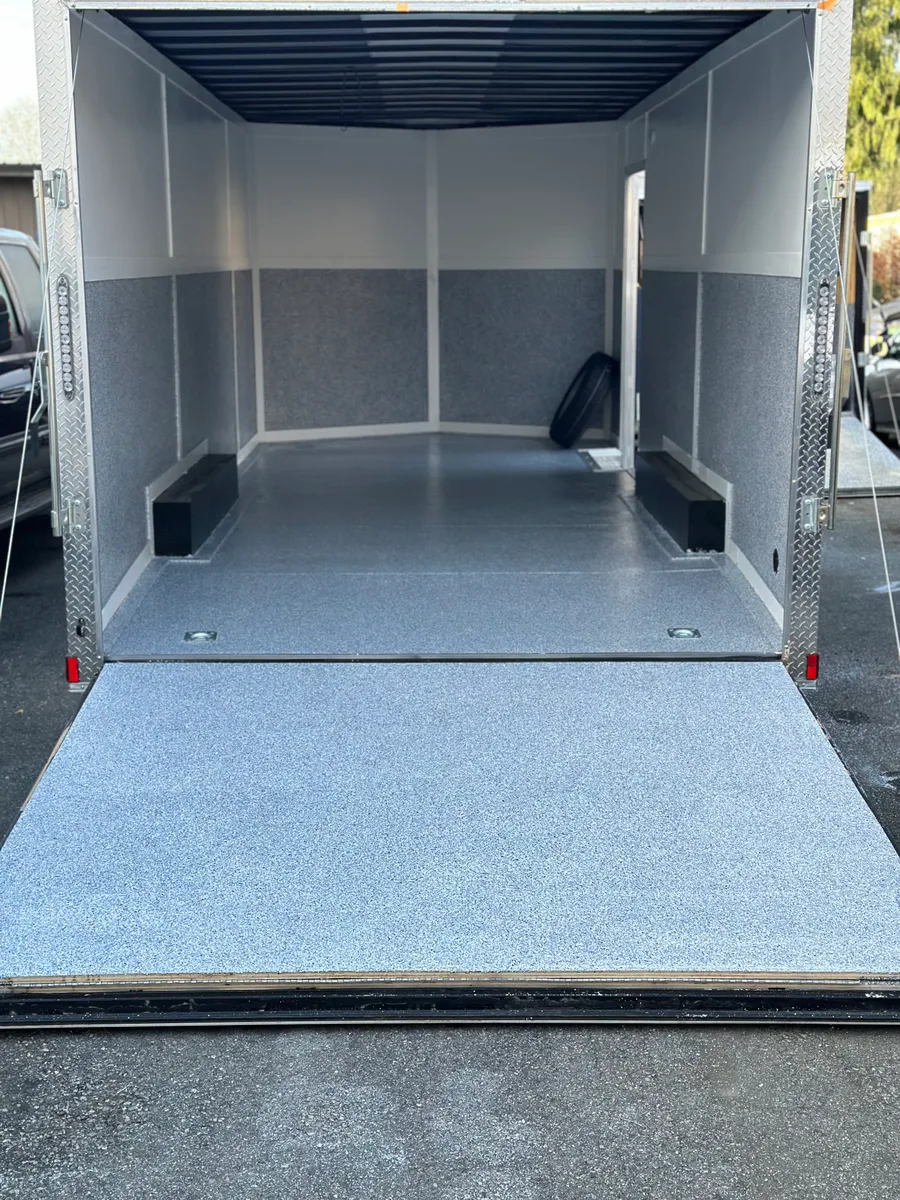 An enclosed car trailer with its ramp down: a grey and white flake coating across the ramp and the full floor, flake panels along the lower walls under white upper walls, and black wheel chocks on each side.