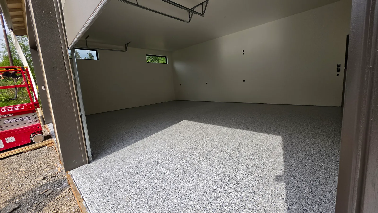 A finished grey and white flake floor coating across an empty new garage, seen through the open overhead door, with white walls, two clerestory windows and a red scissor lift parked outside.