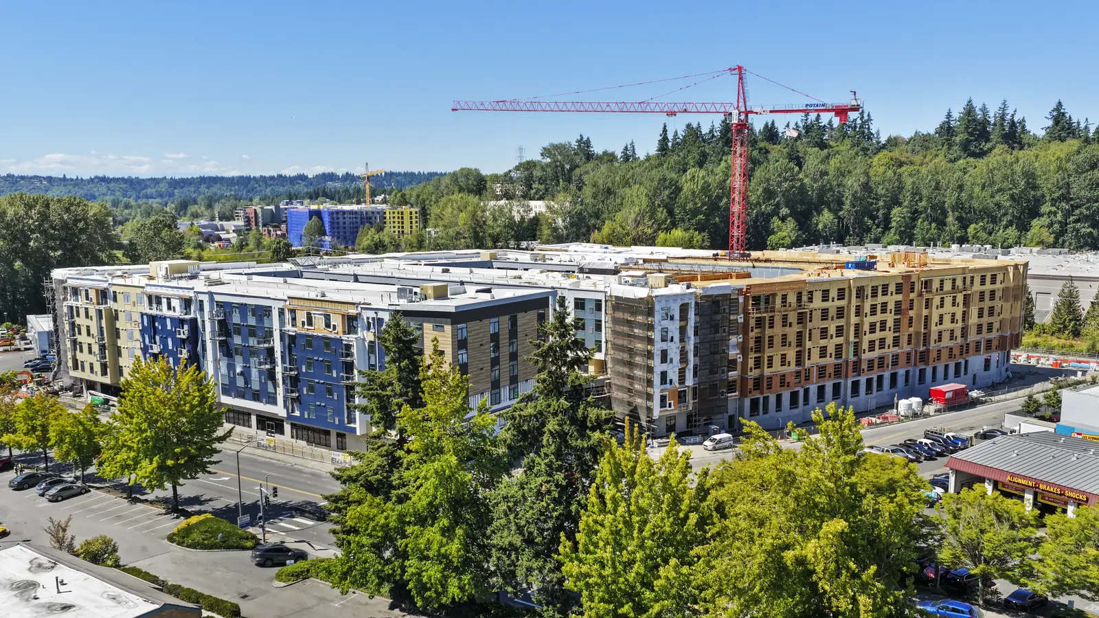 The Foundry at Totem Lake under construction: a six-storey multi-family podium building, part of it clad in blue and tan panels and the rest still in sheathing and wrap, with a tower crane standing over the far end.