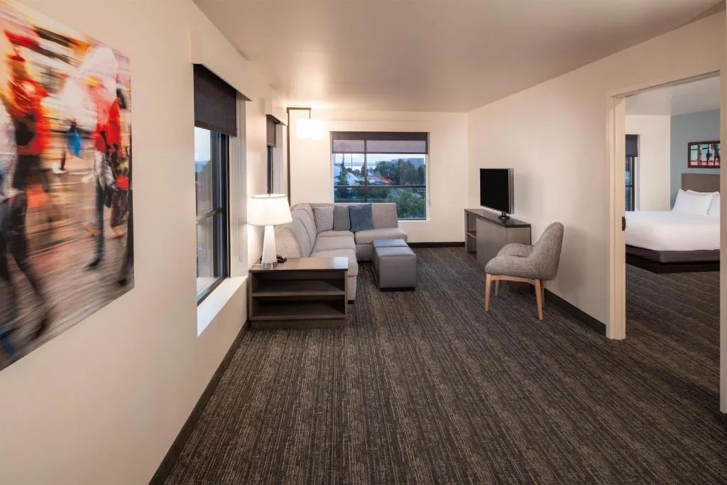 A Hyatt House Seattle suite after the remodel: cream walls and dark baseboards, a grey sectional under a window looking out over the city, a large colourful print on the wall, and the bedroom through an open door.
