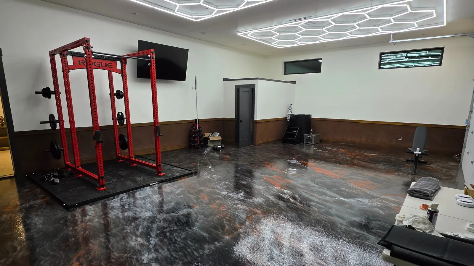 A finished home gym on a mirror-gloss metallic epoxy floor in black, silver and copper, a red squat rack on a rubber platform at left and honeycomb LED ceiling lights reflecting across the floor.