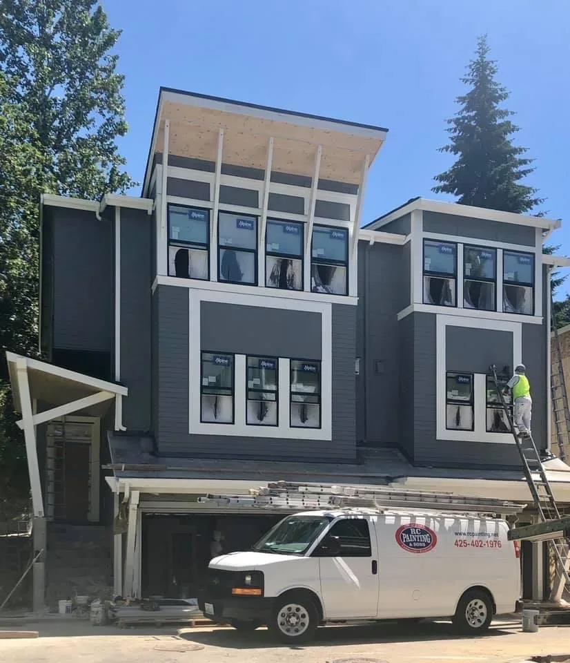 An RC painter on an extension ladder at the upper windows of a new modern home in charcoal lap siding and white trim, stickered windows still in, with an RC Painting & Sons van parked in the drive below.