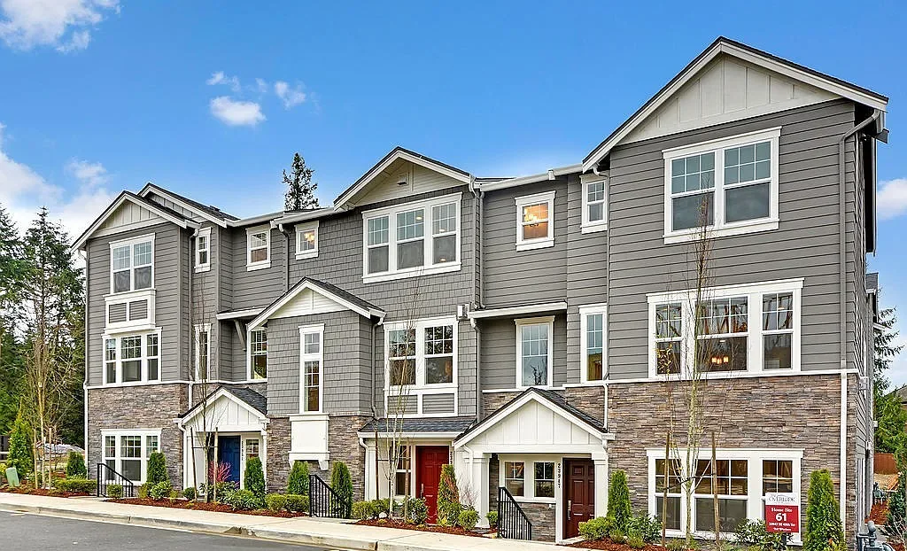 Three-storey townhomes at the Ridge at Big Rock in Duvall: grey lap and shingle siding with white trim, stone bases, and red and blue front doors along a new sidewalk.