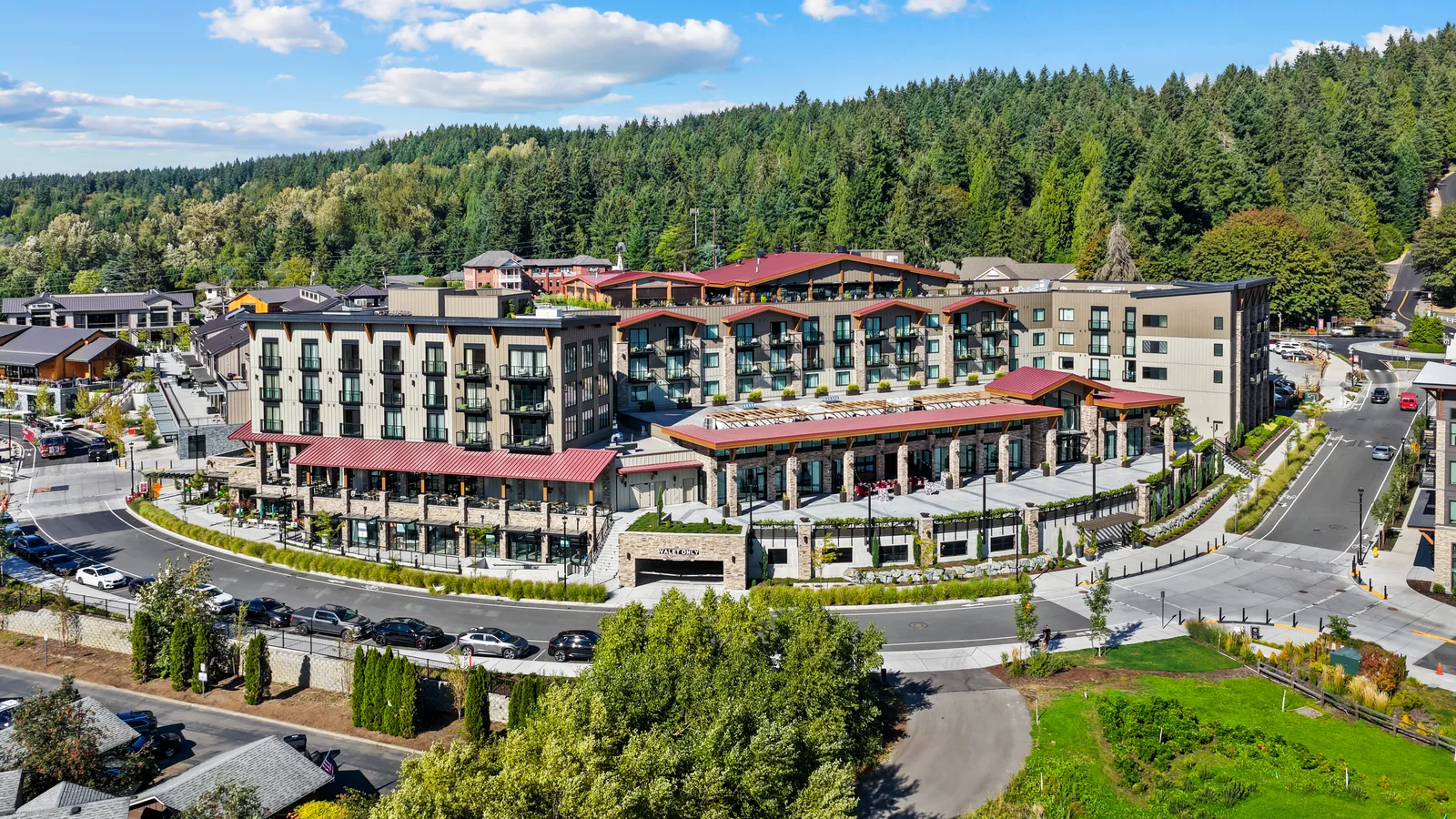 The back of the Somm Hotel in Woodinville from the air: a four-storey hotel in pale board-and-batten siding with dark window trim and balconies, red standing-seam roofs over stone-columned colonnades and terraces at street level, a valet entry on the curved road below, and forested hills rising behind.