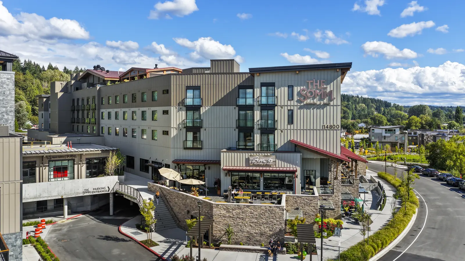 The Somm Hotel in Woodinville: a five-storey hotel clad in grey and tan vertical metal siding, THE SOMM lettering on the gable end, with the Bin 41 restaurant and its red awnings at street level above a stone base.