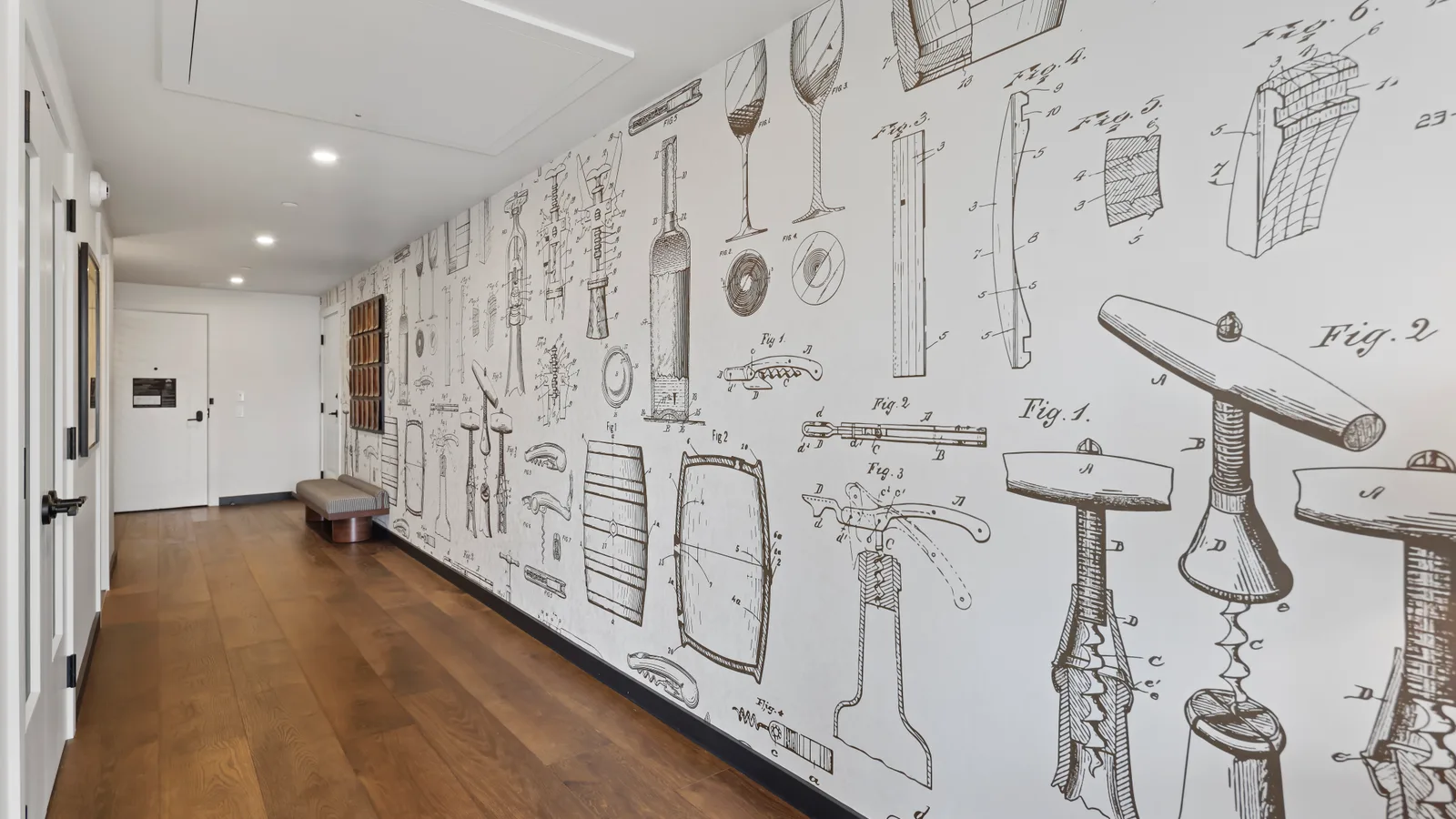 A corridor at the Somm Hotel papered floor-to-ceiling with line drawings of corkscrews, bottle openers and barrels in the style of patent illustrations, running the length of a white wall above a dark timber floor.