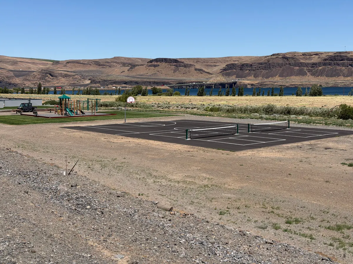 A new black sport court with white pickleball lines and two nets, a basketball hoop at one end, beside a playground on open dry ground, with a river and basalt cliffs beyond.