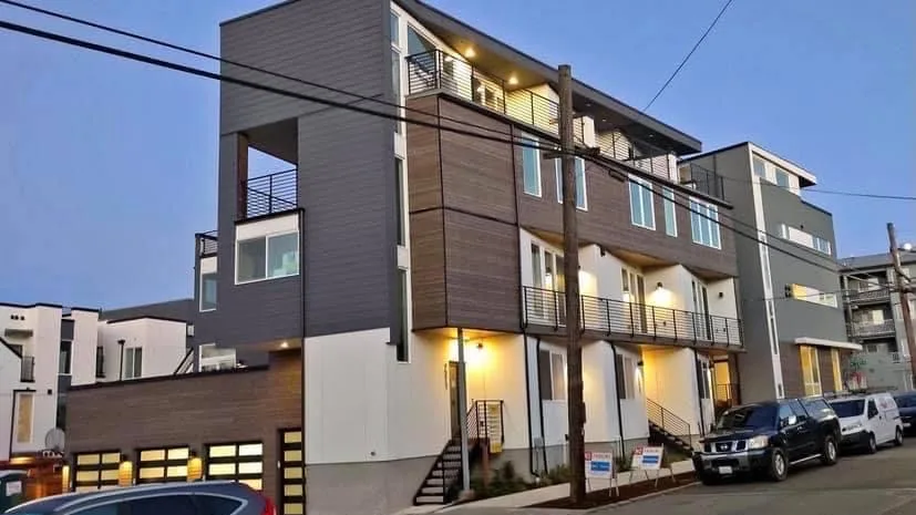 A four-storey modern townhome building at dusk: grey and wood-toned lap siding over white panels, black railed balconies, and warm lights glowing at each entry along the street.