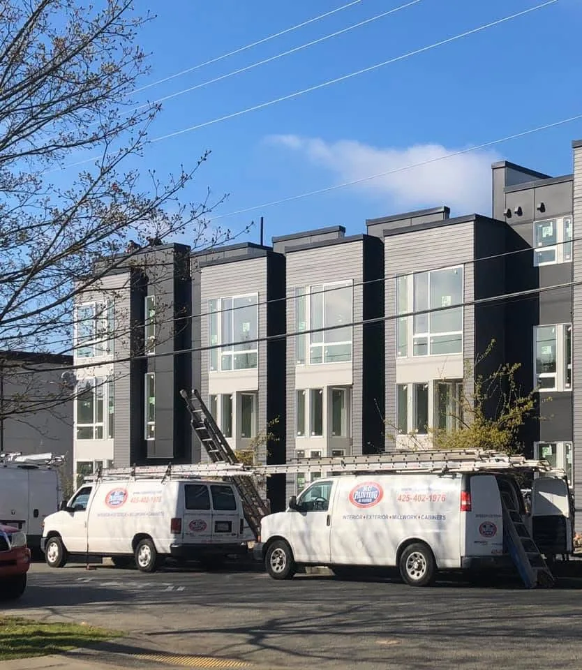 Two white RC Painting & Sons vans with ladders on their roof racks, parked in front of a row of new four-storey townhomes clad in grey and charcoal siding with tall white-framed windows, under a clear blue sky.