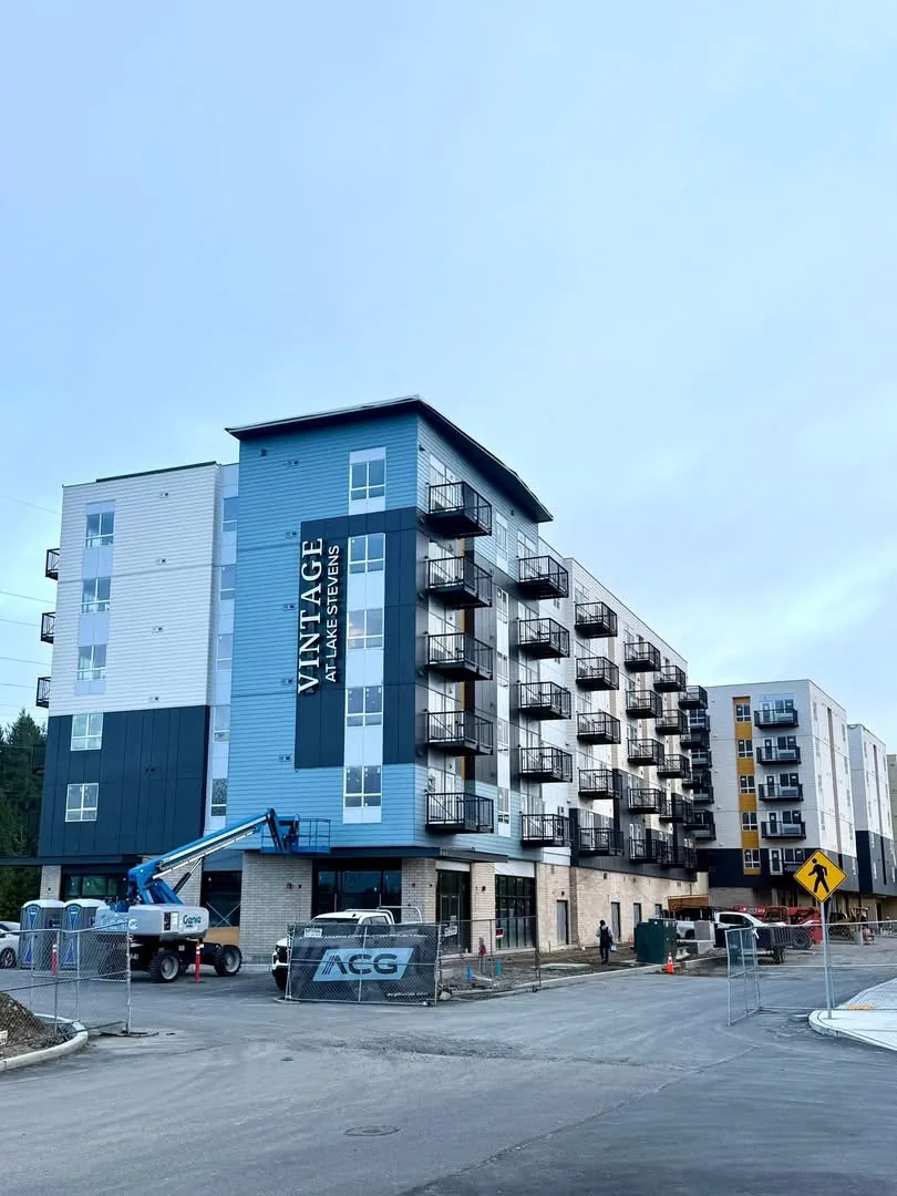 Vintage at Lake Stevens under construction: a five-storey apartment building in blue and white siding with navy panels and black balconies, its name in white on a tall navy panel, a boom lift and site fencing out front.