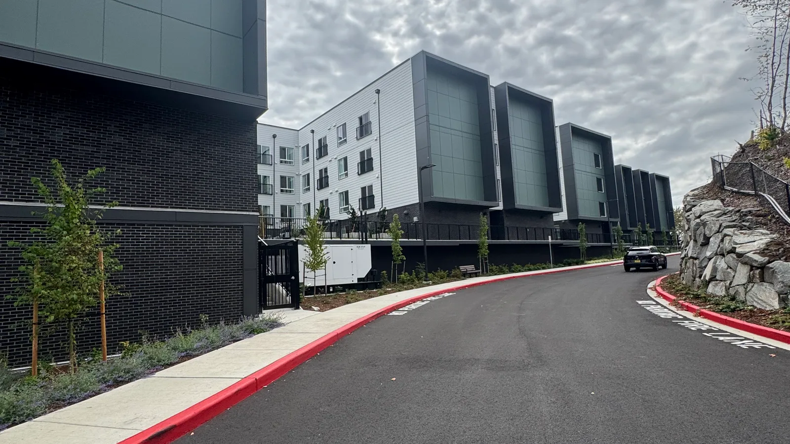 Vintage at the Overlook from its curving entry drive: white lap-sided wings with deep grey-green panelled bays over a dark brick base, red-painted curbs along new asphalt, and a rockery and young trees lining the road.