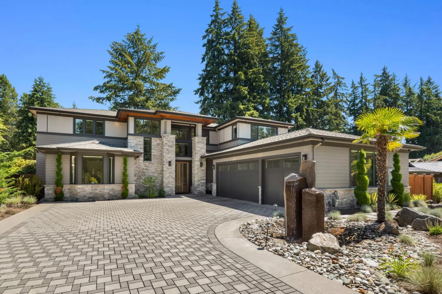 A two-storey Northwest contemporary custom home in Bellevue: grey lap siding with dark trim, a stacked-stone entry tower, cedar soffits under low hipped roofs, a charcoal two-car garage, and a paver drive curving past a rock garden under tall firs.