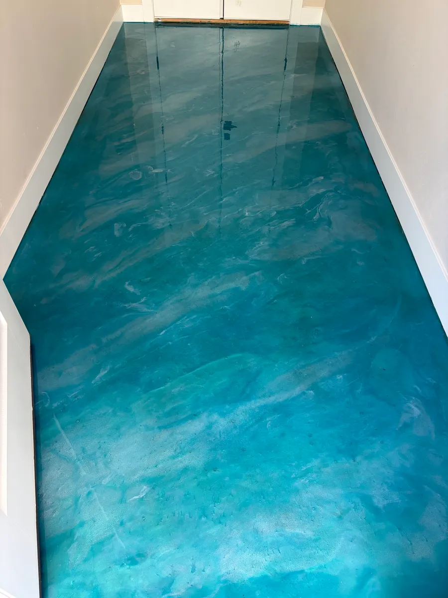 A finished metallic epoxy floor running the length of a narrow hallway: glossy teal swirled with white and pale blue, like water, between white walls and baseboards.