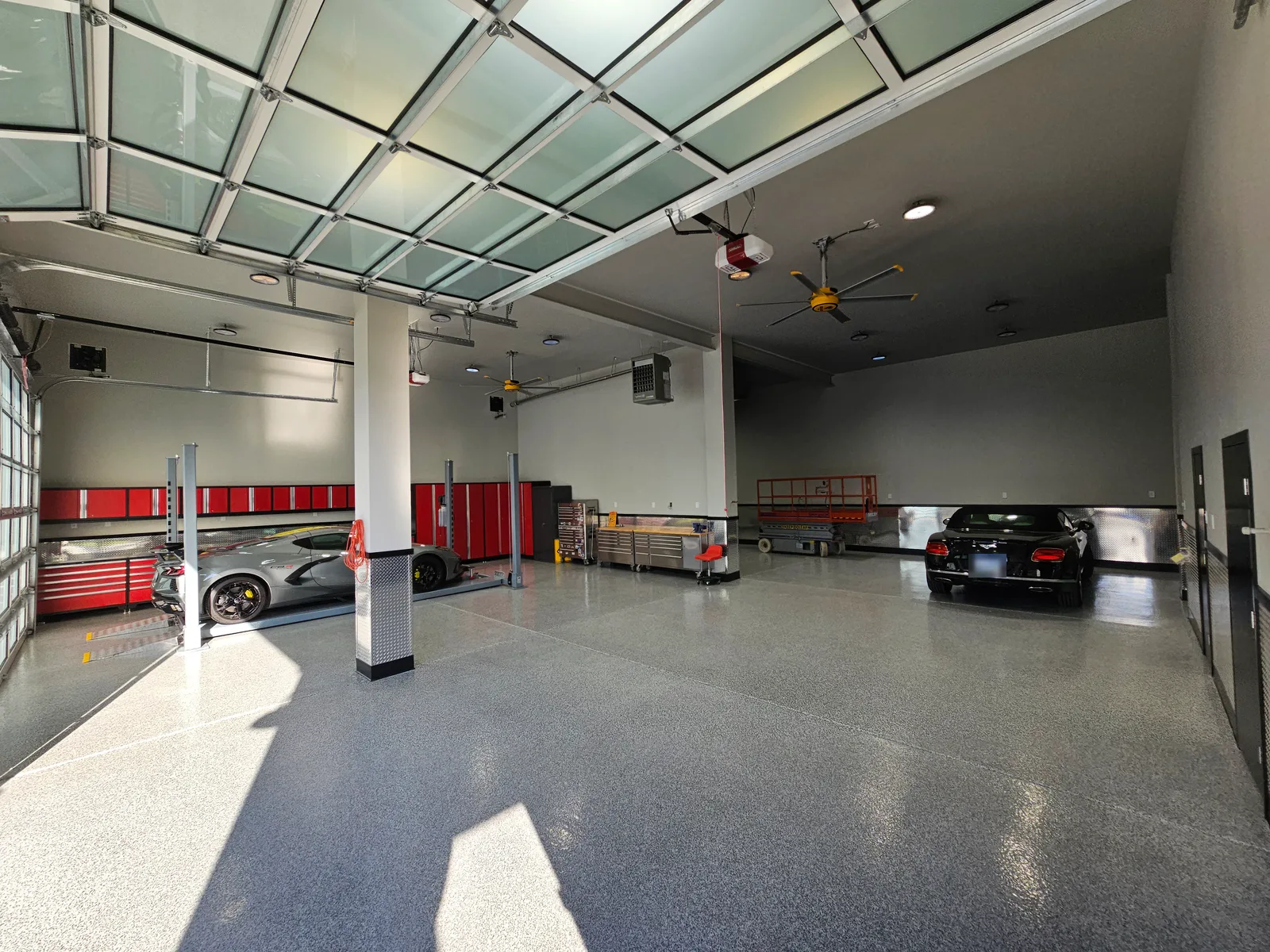 The same garage after coating: a glossy grey flake floor reflecting the skylight doors, a sports car on a lift beside red tool cabinets at left, and a black convertible parked at right.