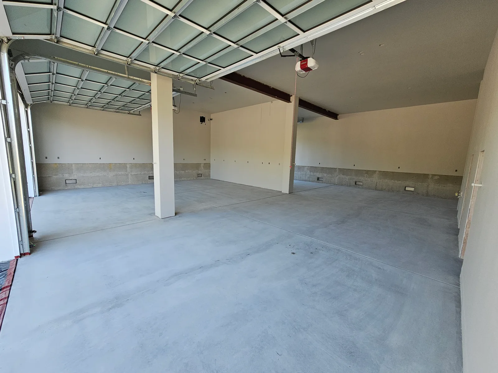 A large garage before coating: a bare grey concrete slab with saw-cut control joints, a white column between the bays, and unfinished board-formed concrete along the base of the walls.