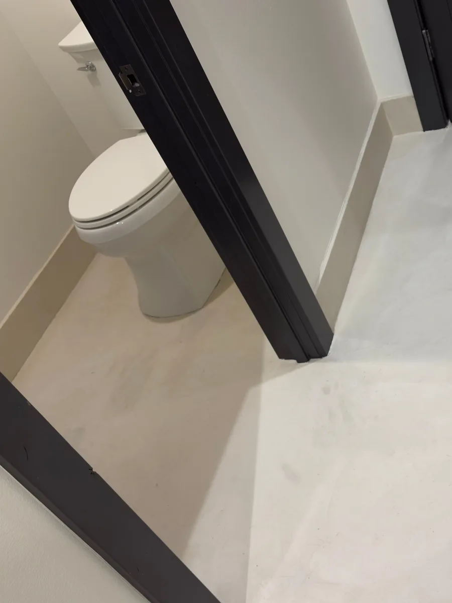 A finished microcement bathroom floor in pale warm grey, one seamless surface running from the vanity area through to the toilet room, framed by black door casings and cream baseboards.
