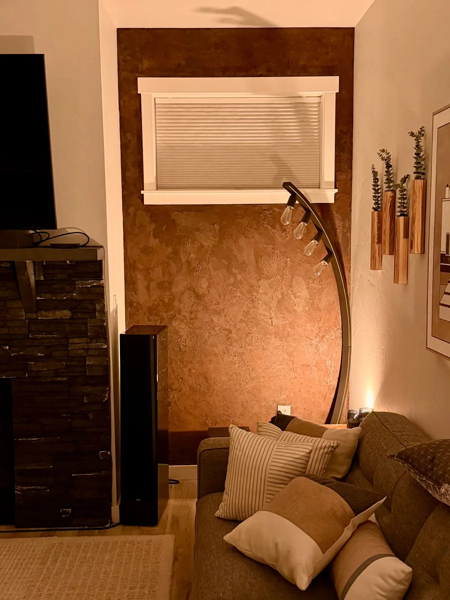 A living-room accent wall finished in a mottled, trowel-textured microcement with a rusted iron look, warm copper and brown tones catching the lamplight around a white-trimmed window.